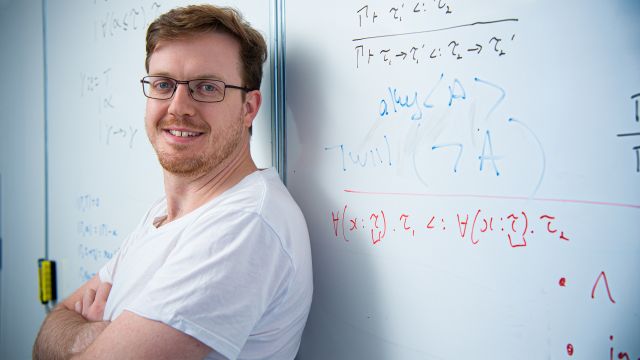 Smiling man stands in front of a whiteboard showing mathematical equations.