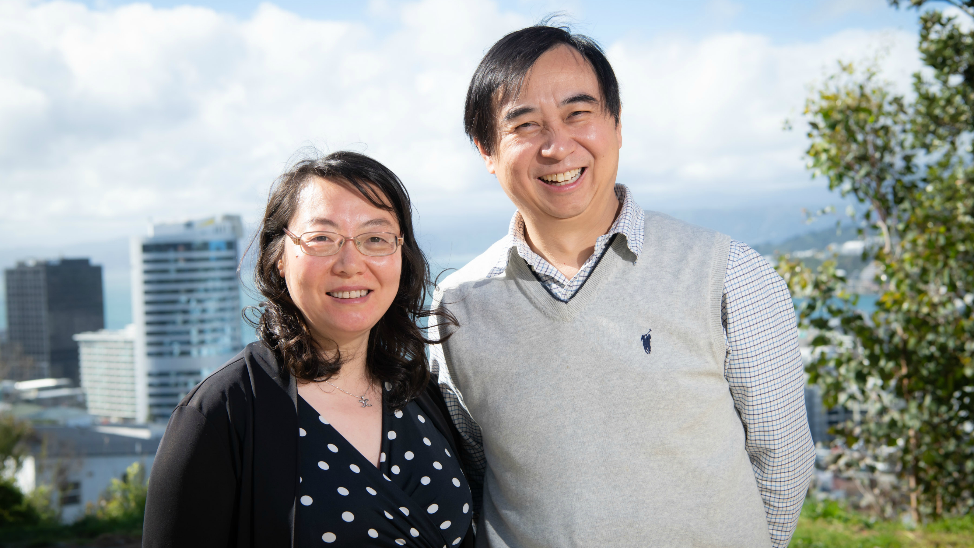 Husband and wife stand smiling in front of a harbour view.