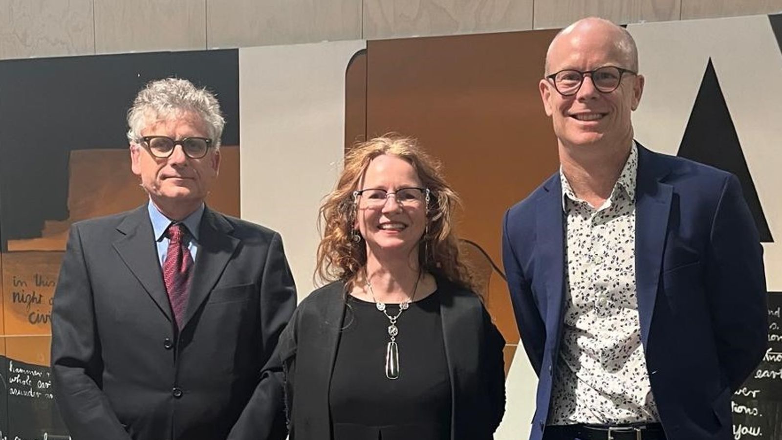 Three people stand side by side, smiling in front of a modern art backdrop.