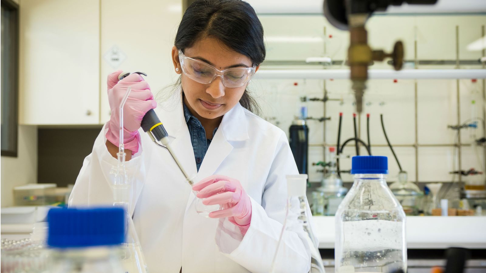 A woman in a lab coat holds a pipette and round based transparent flask while conducting an experiment.