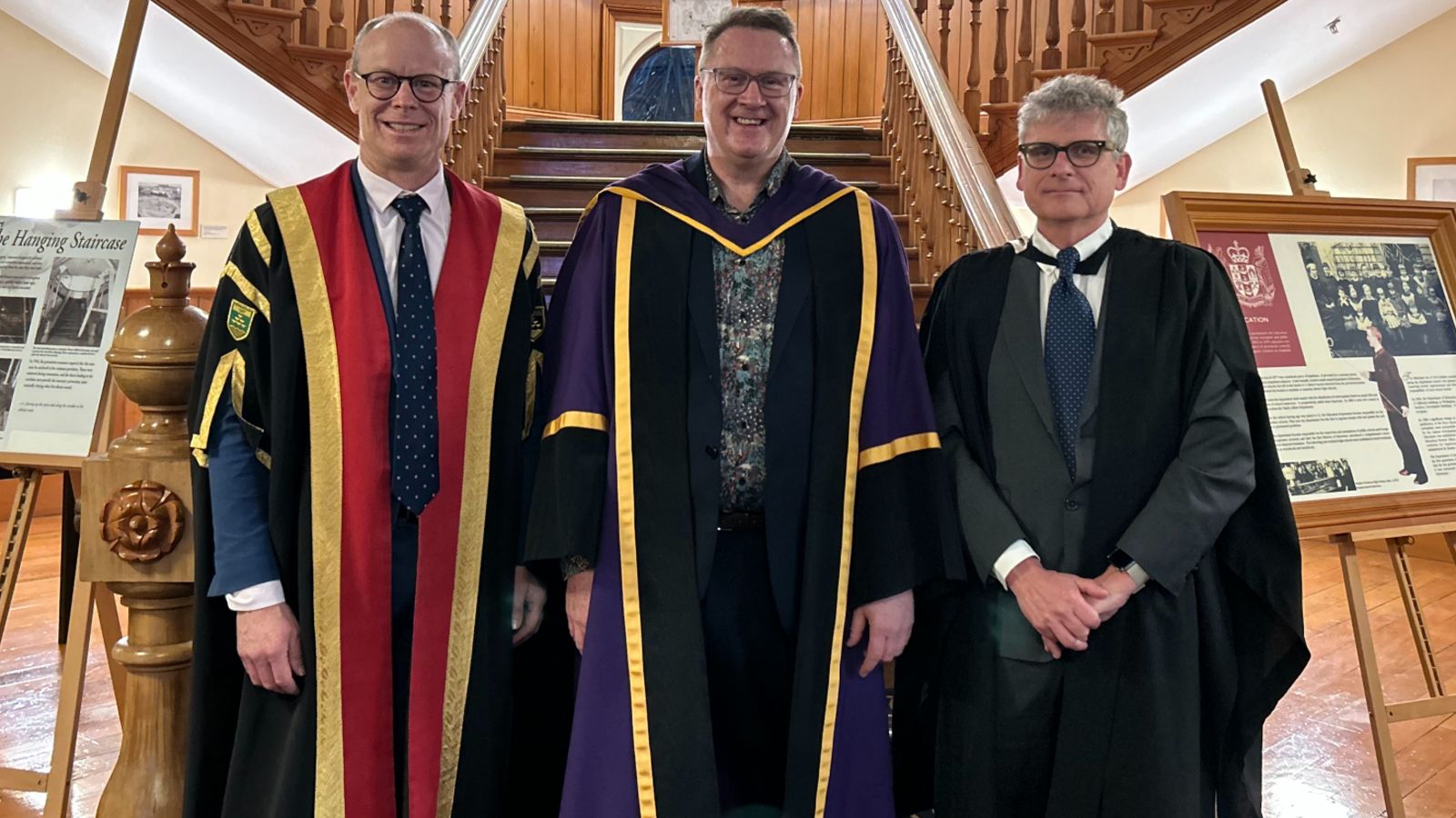 Three men dressed in academic regalia stand in front of a staircase in a wood-paneled hall.