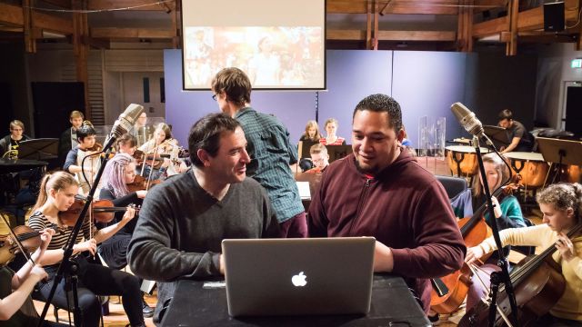 Music film scoring – A professor and a student look at a laptop, and stand between two freestanding microphones, while others play orchestral instruments in the background.