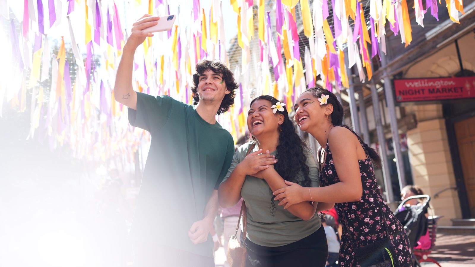 Three students take a selfie at the Cuba Dupa street parade, with white, purple and orange streamers behind them..