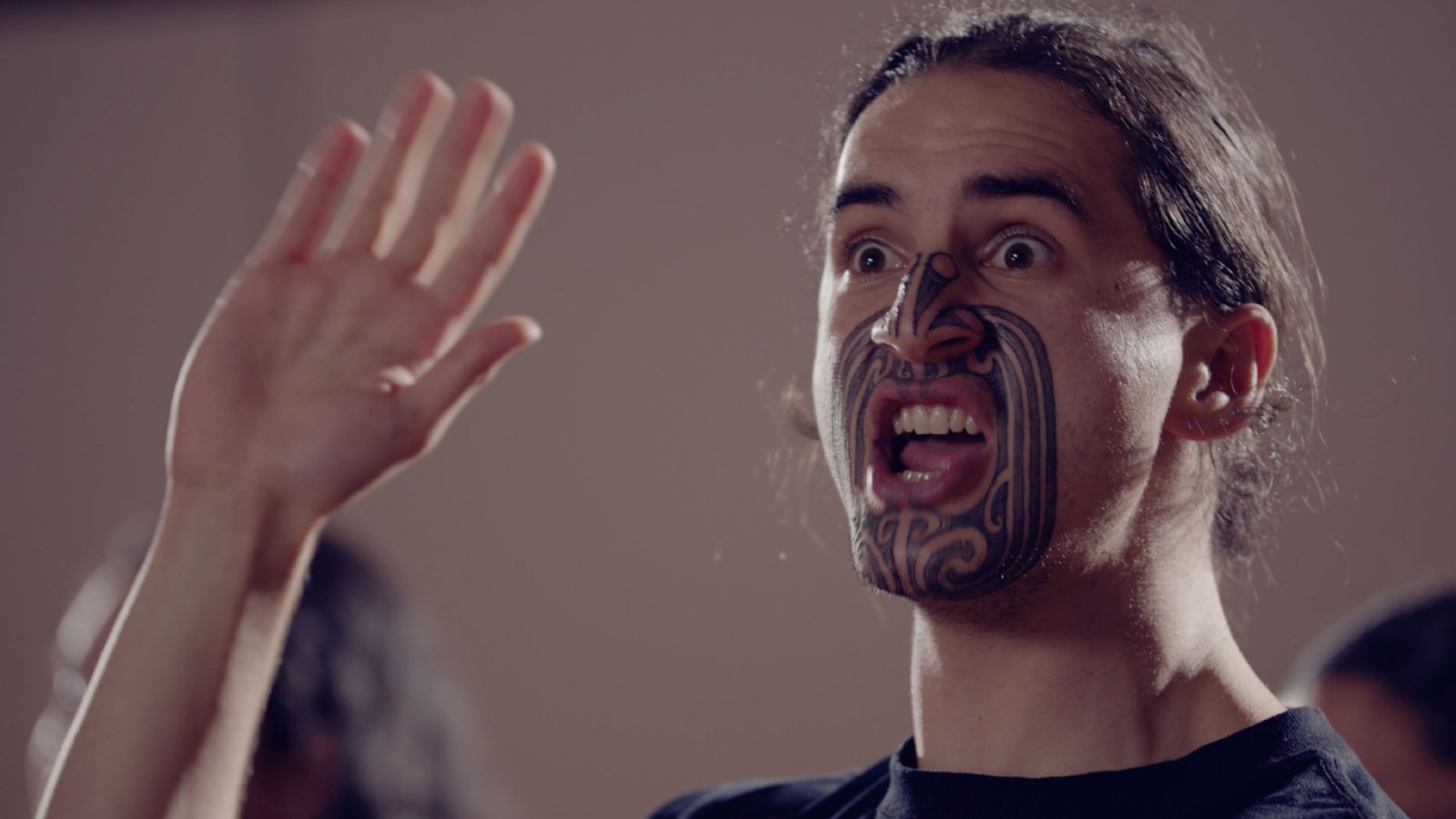 A close up of a man's face, which is partially tatooed with an indigenous design. He is holding one hand up and his expression is of surprise or fierceness.