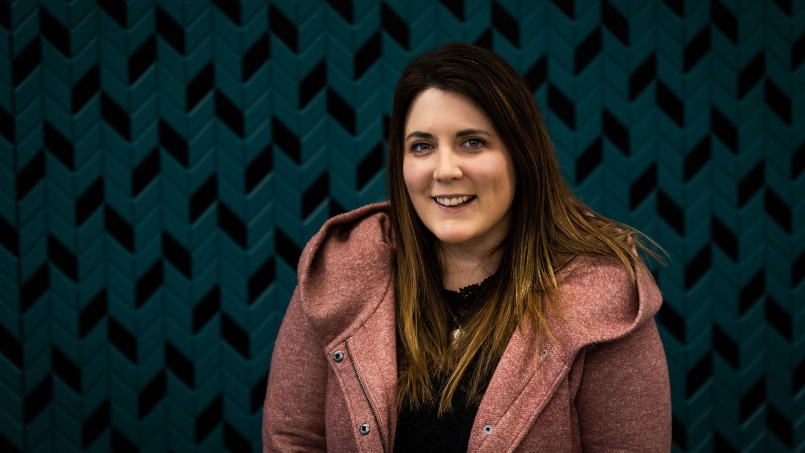 Portrait of Lisa McLaren wearing a pink jacket in front of a teal chevron patterned wall. 