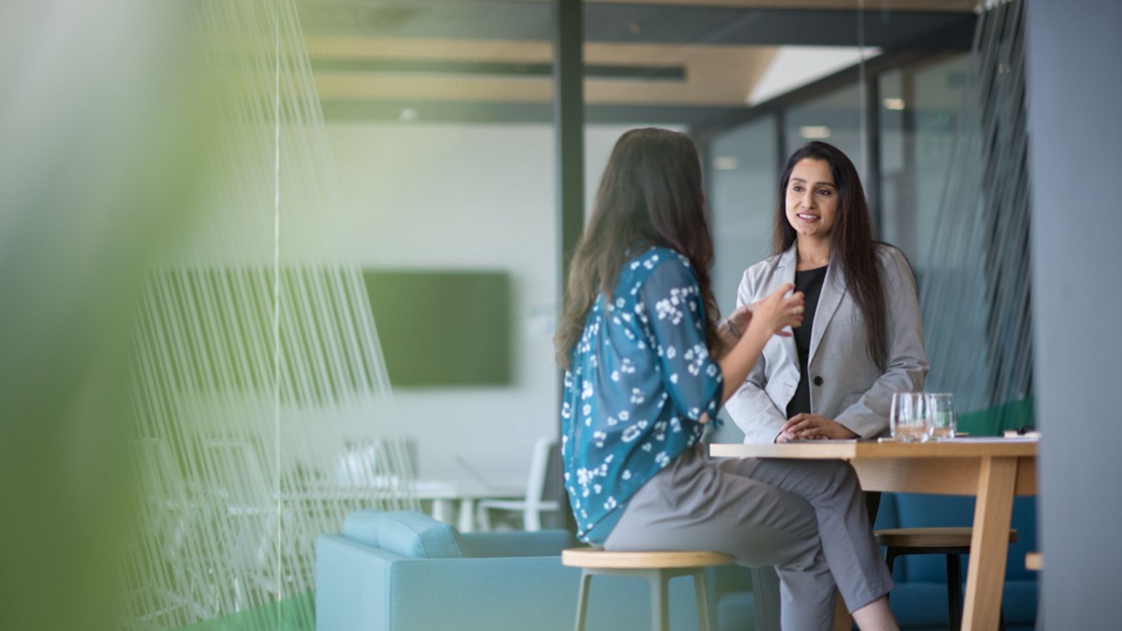 Two female postgraduate students with long dark hair having a conversation at a table in a corporate setting.