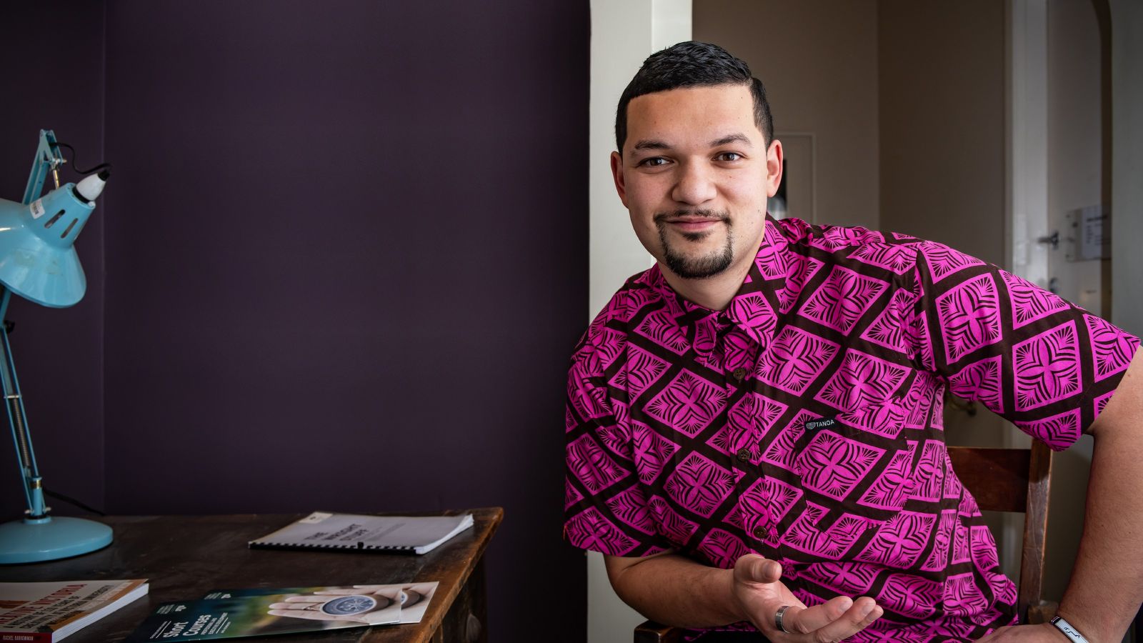 Image of Leki Jackson-Bourke wearing a pink shirt with a Polynesian pattern next to a desk with a Te Herenga Waka—Victoria University of Wellington Short courses booklet on it. 