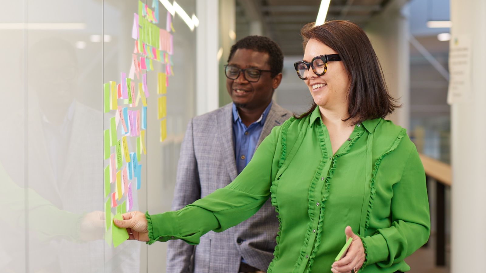 A smiling woman in a green blouse and a man in a suit stand in front of a whiteboard covered with colourful post-it notes