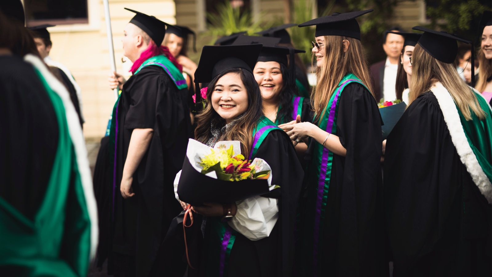 An image of an Asian woman smiling amongst a group of graduates wearing caps and gowns at their graduation parade.