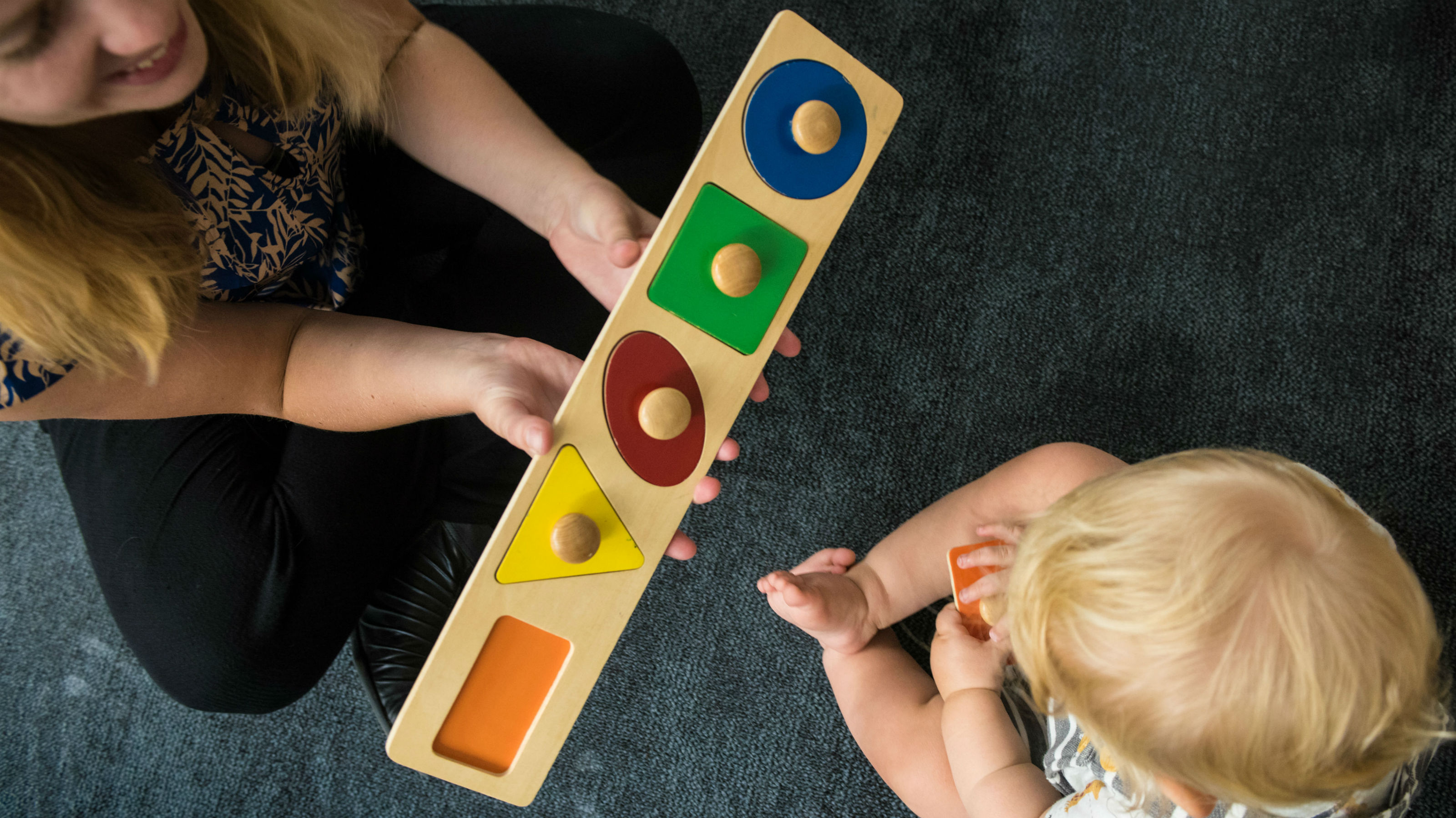 A view from above, a woman holds a wooden puzzle of four shapes, a young child holds an orange rectangular piece in his hand.