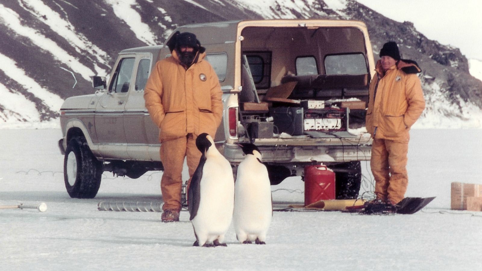 Two men in jumpsuits in Antarctica with penguins in foreground.