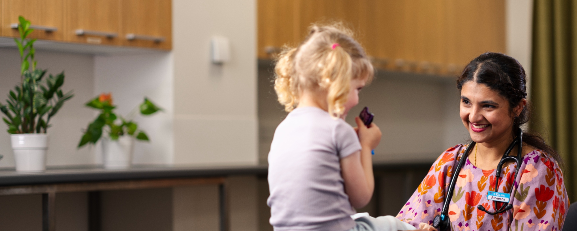 A nurse in colourful scrubs with a stethoscope around her neck smiles at a young girl.