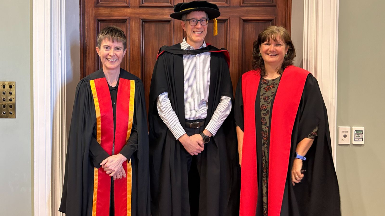 Three people in academic robes stand in front of a wooden panelled door.