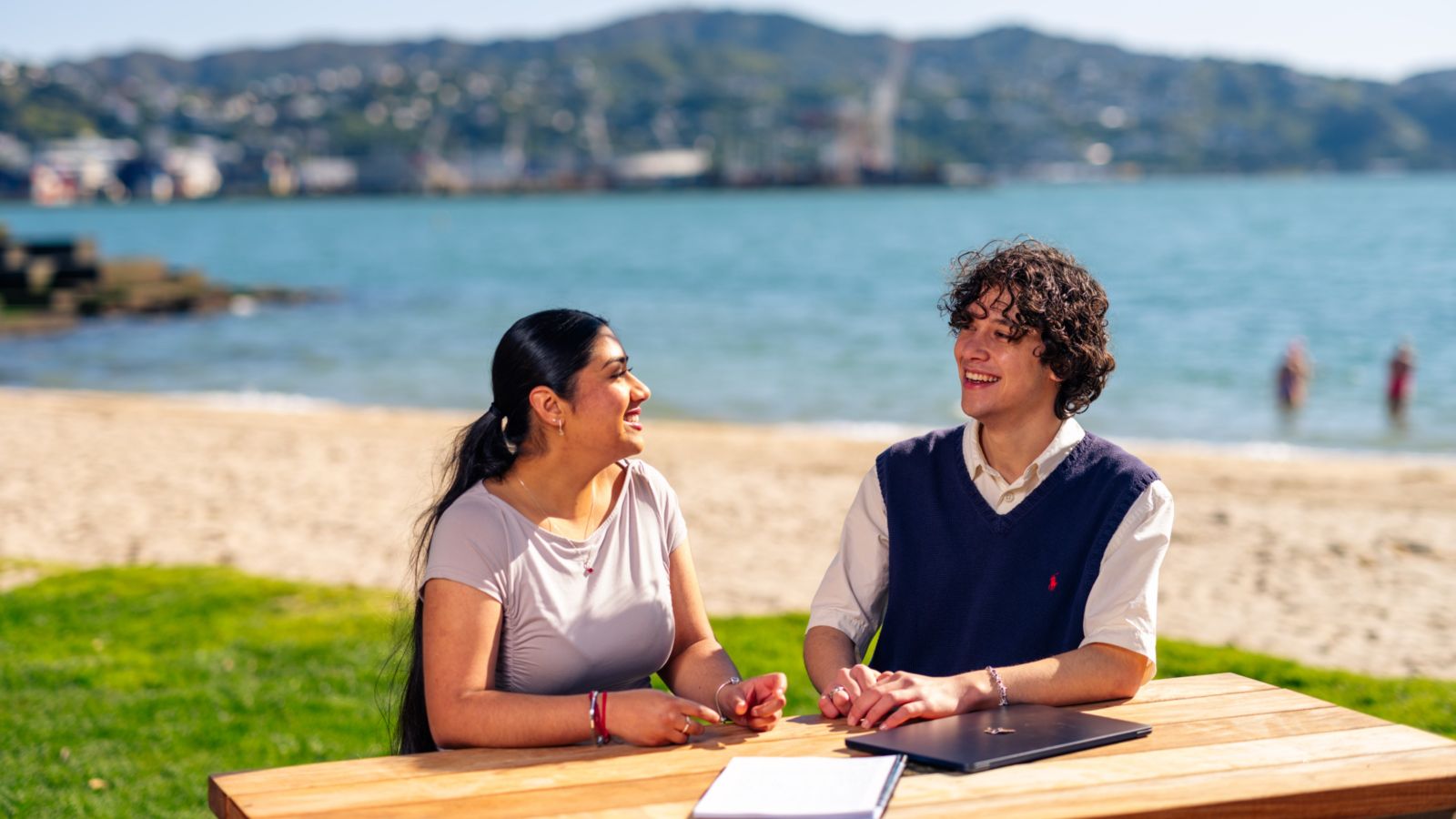 A male and a female student sit at a picnic table in Wellington's Oriental Bay, with a laptop and notebook in front of them.