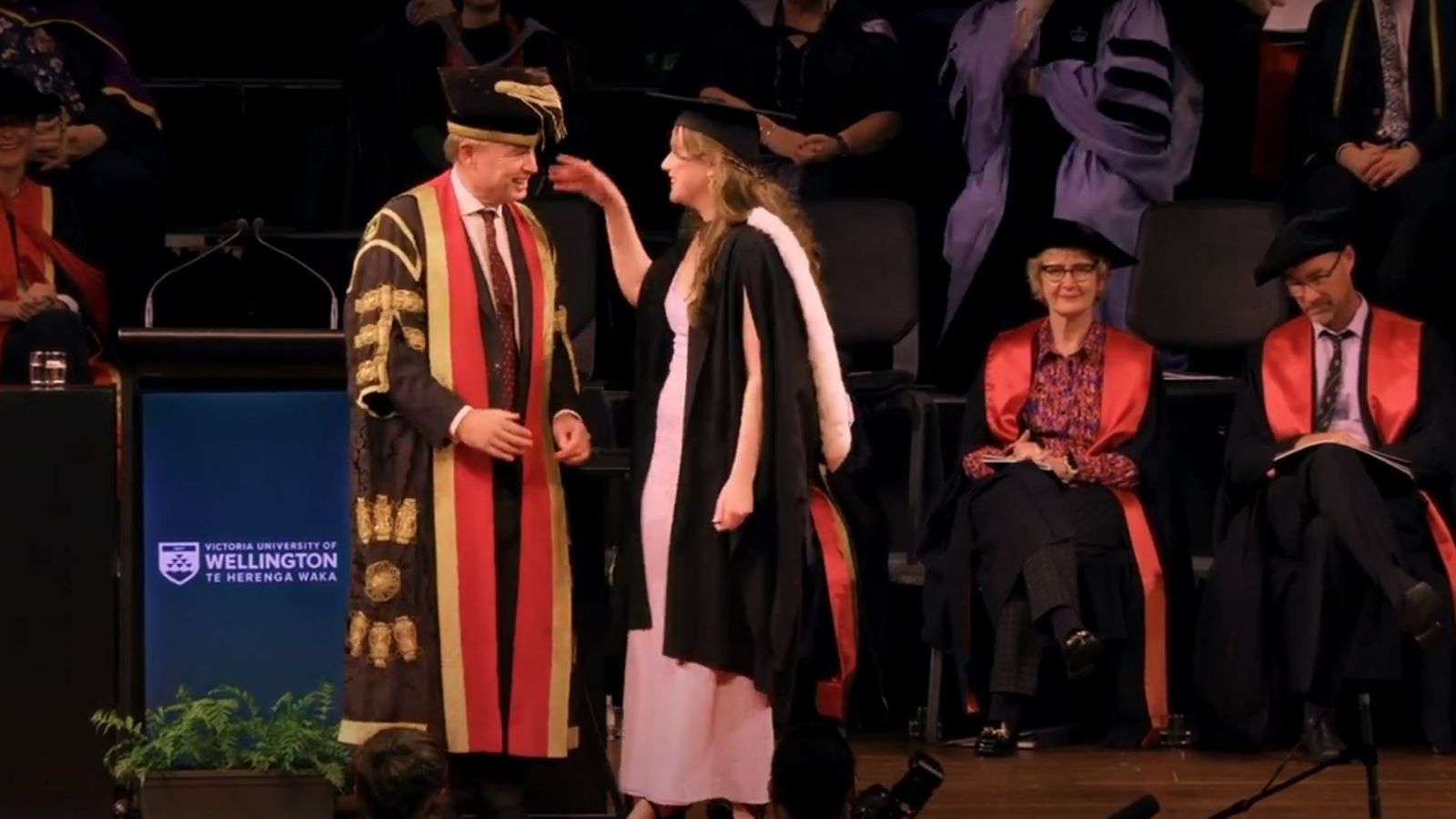 A woman dressed in academic robes reaching up toward the Chancellor