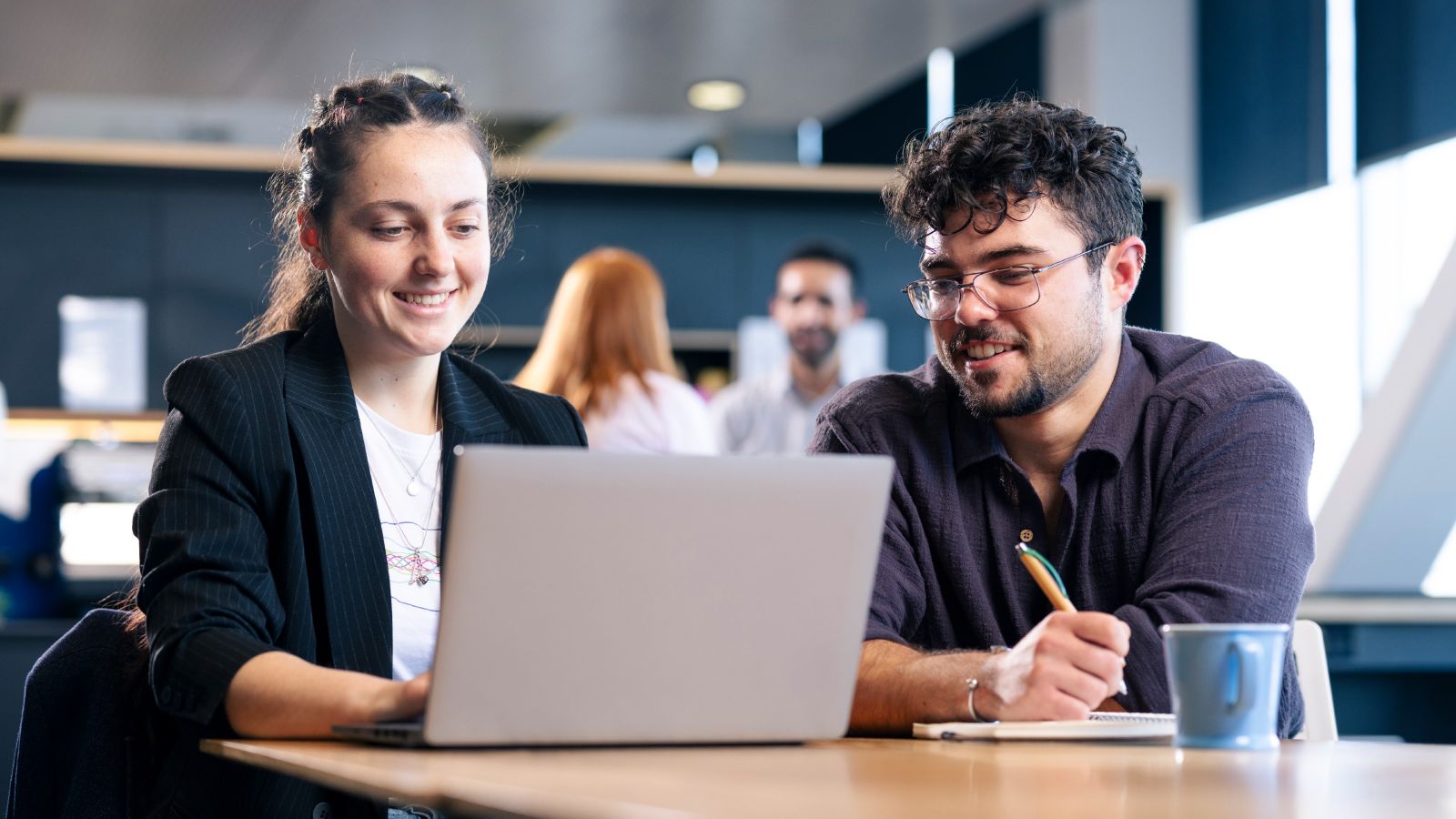 Two students sit at a table with a laptop in front of them.