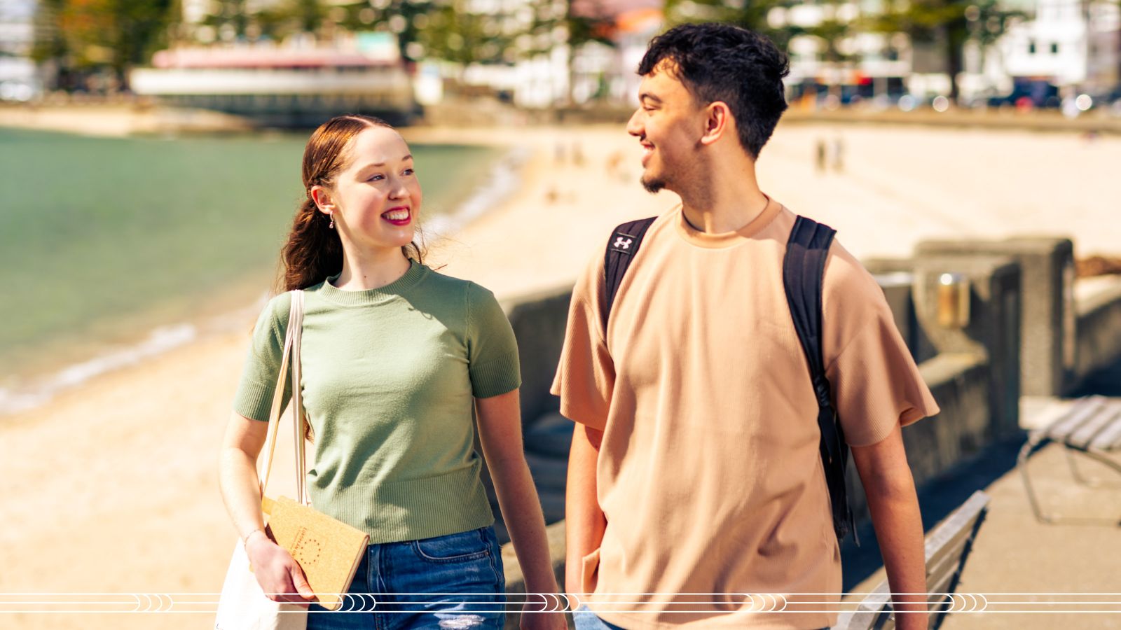 A female and a male student are walking along, smiling at each other, with a city beach scene out of focus in the background