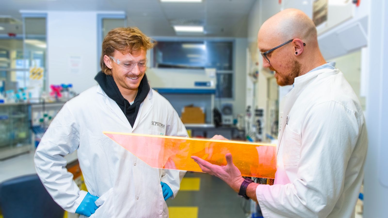 Two researchers in white lab coats talking with each other, one is holding a sheet of orange luminescent material.