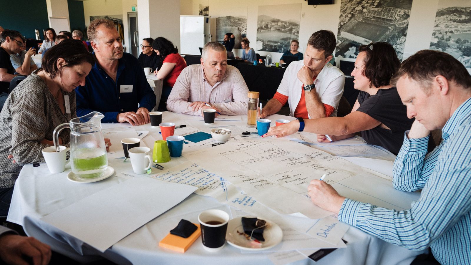 Group of researchers seated at a round table filled with cups, papers, and pens talking with each other.