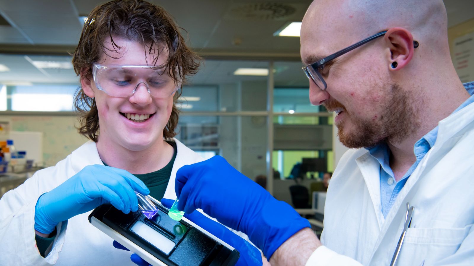 Two researchers hold luminescent matter in small containers up to electronic tool.