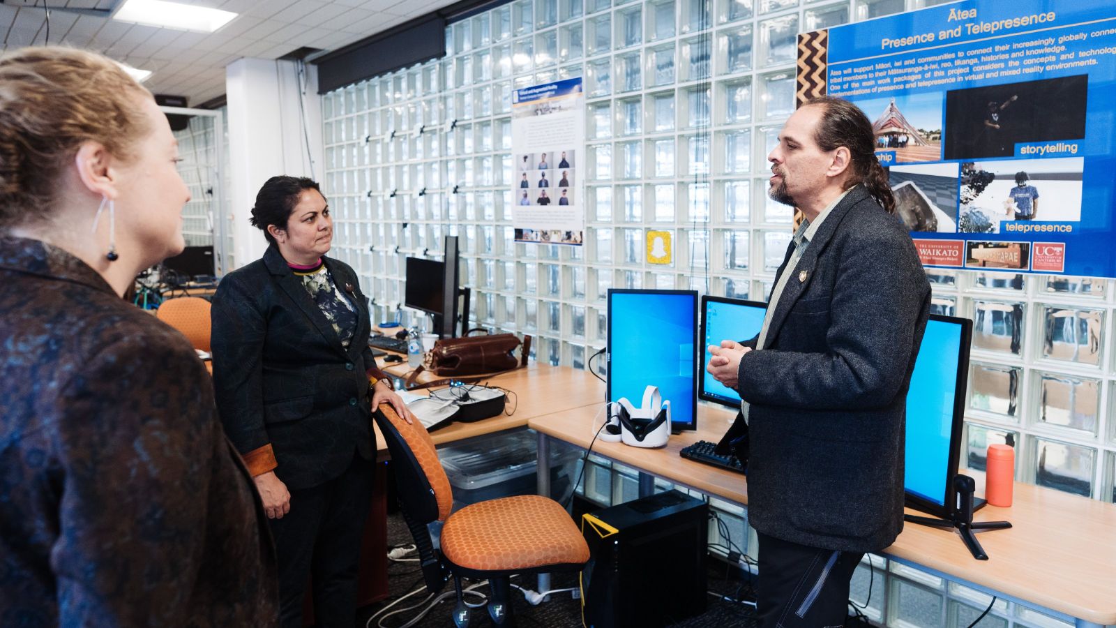 Two people listening to a researcher present while standing infront of a sign and computer screens with a virtual reality headset pugged in to it.