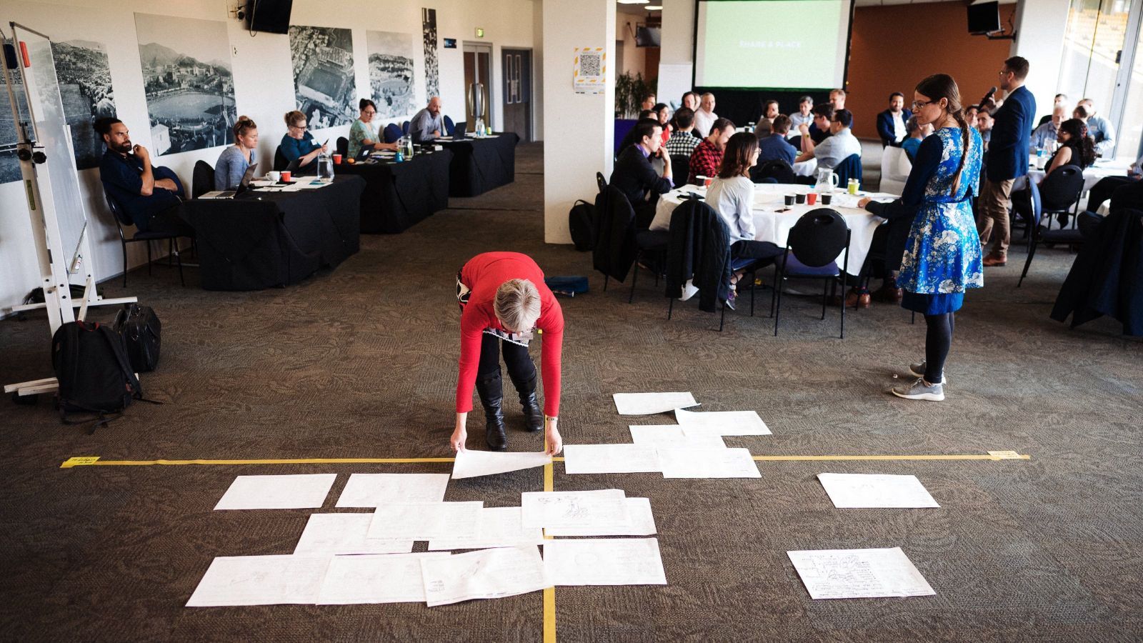 Workshop attendee places a piece of paper among other pieces paper arrange in a yellow axis on a carpeted floor with other attendes seated and standing in the background.