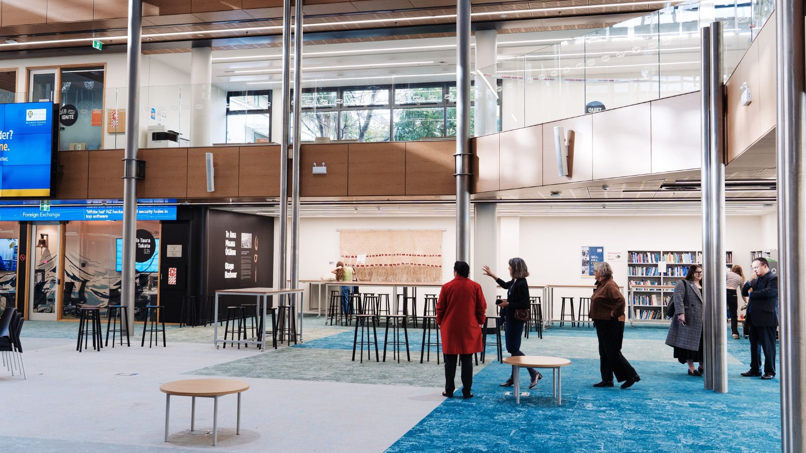 Ground floor of University of Otago library with scattered furniture, large columns, multi-coloured carpet, and small groups of people standing and talking to each other.