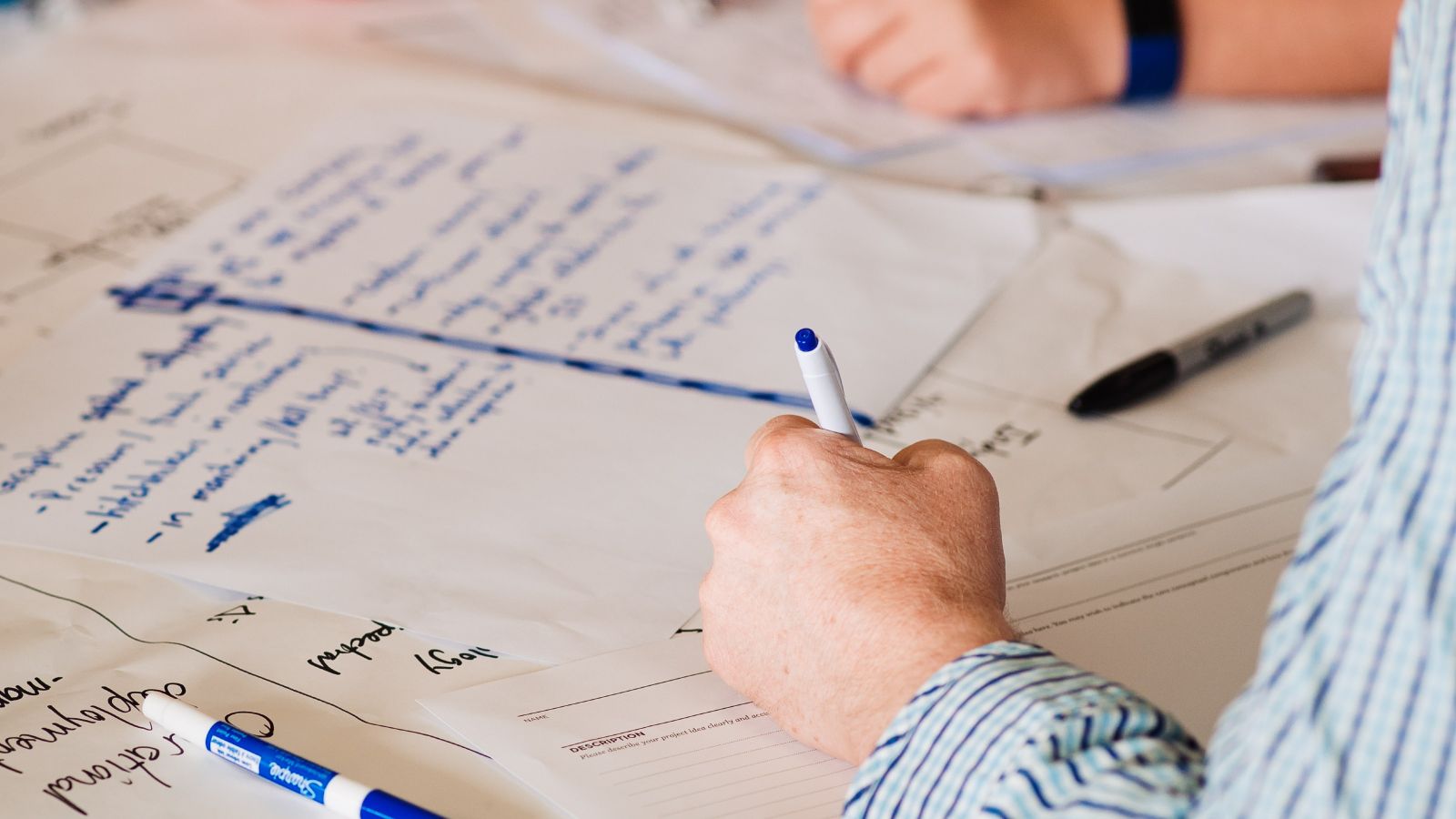 Photo of a A3 paper with blue writing and a hand hovering over the page holding a pen.