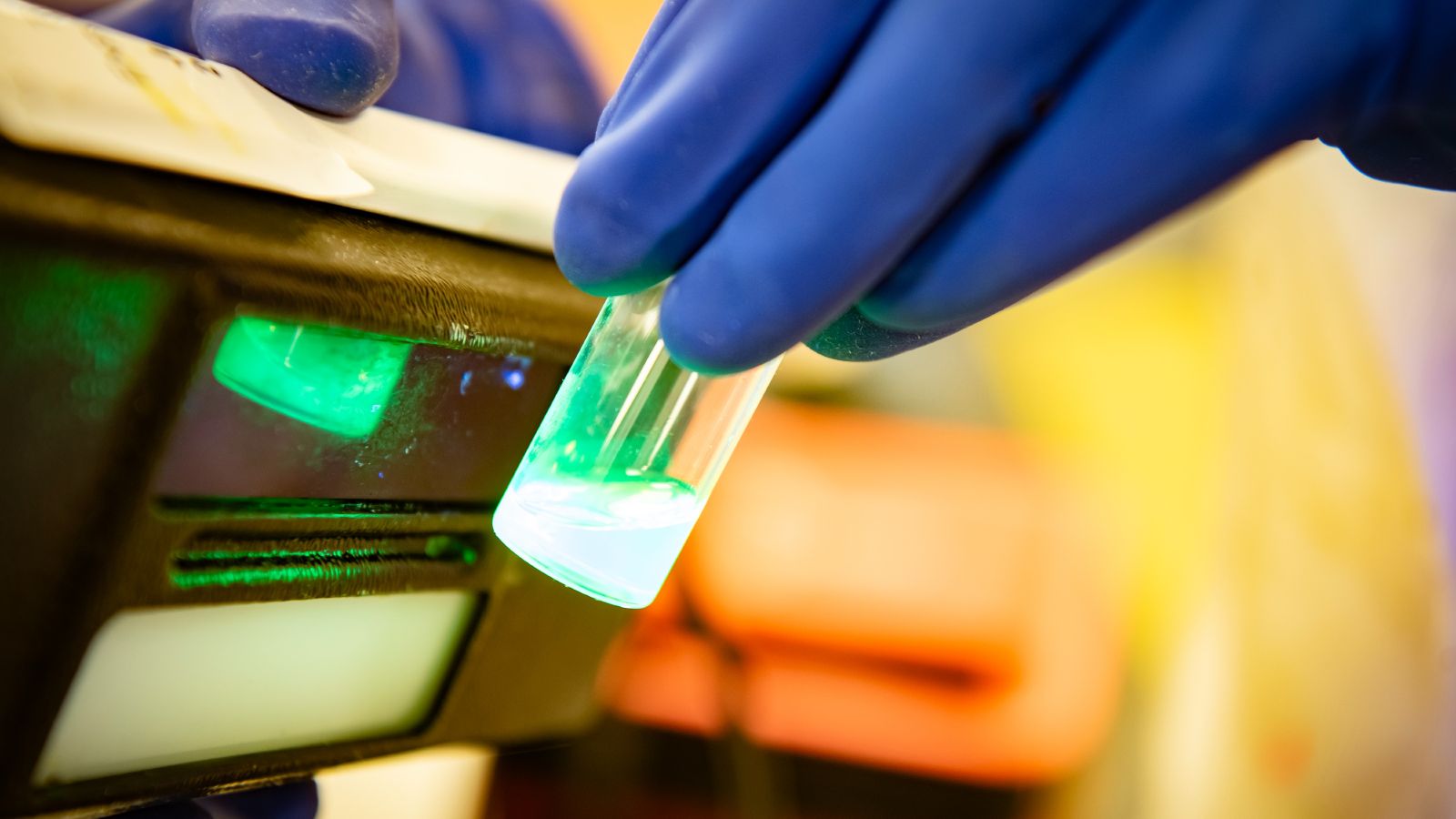 Bright green luminescent material in a small bottle held by a hand wearing blue rubber gloves