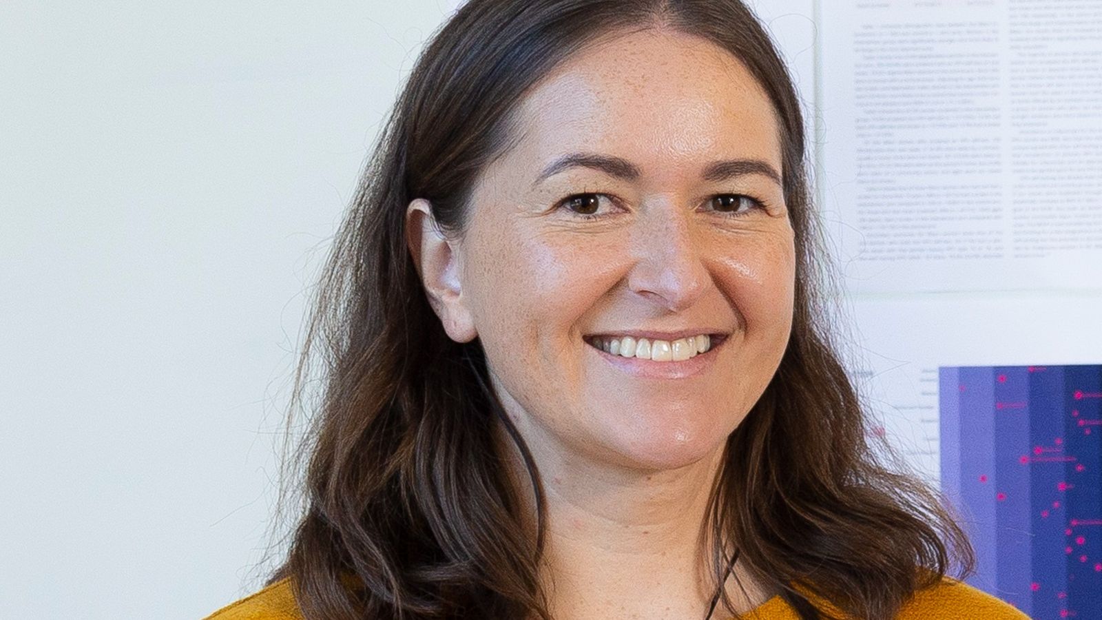 Portrait of Anna Adcock smiling at the camera. She has brown hair and wears a yellow top.