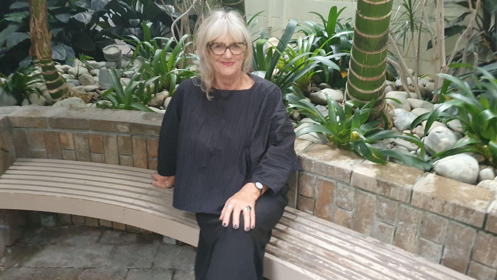 Portrait of Bronwyn McGovern. Bronwyn has light hair and wears black and is seated amongst greenery.