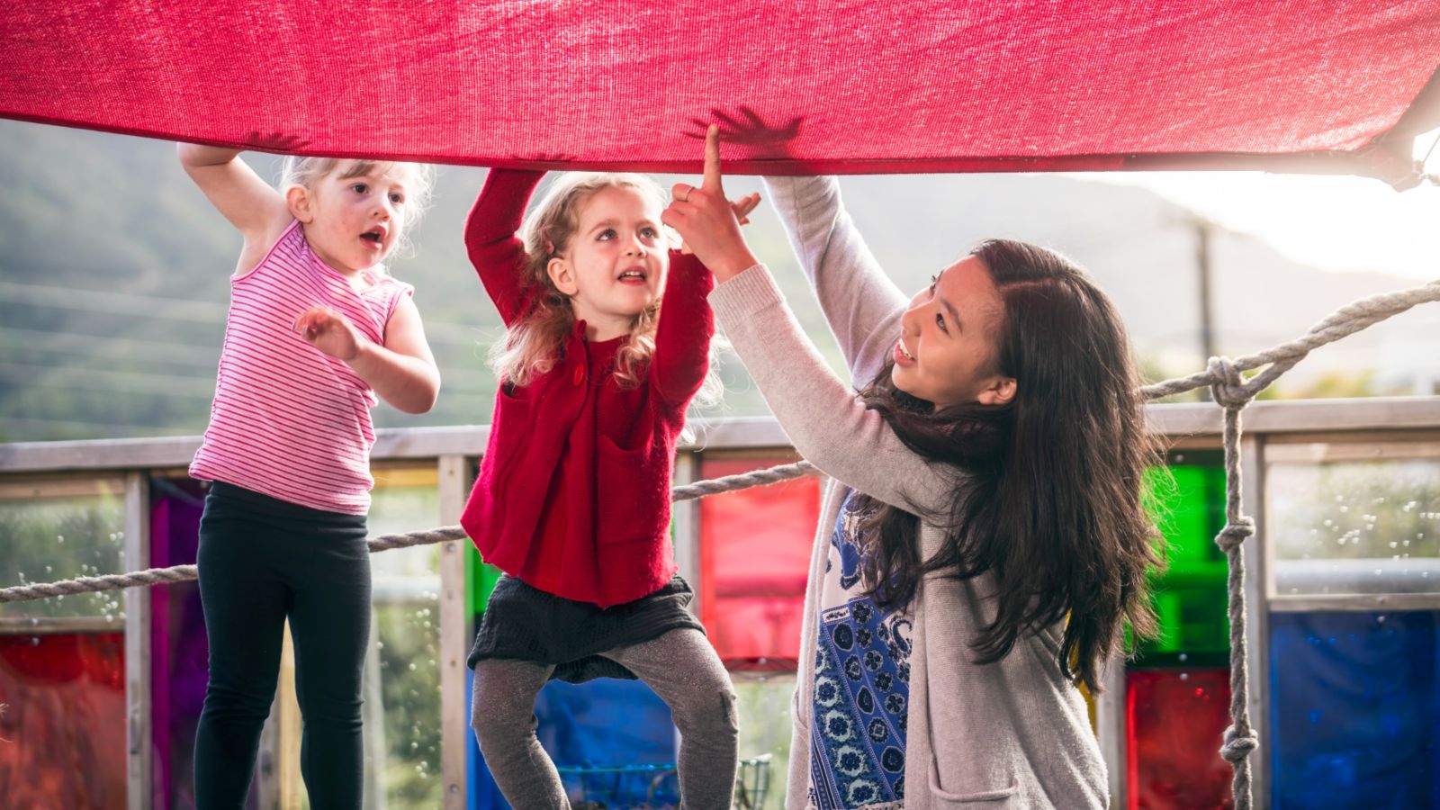 Female ECE teacher showing two preschool girls hand shadows through a red shade sail.