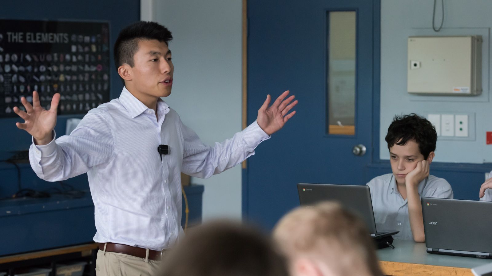 Male secondary teacher gesturing with his arms teaching science with boys in background on computers