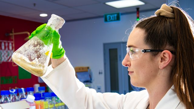 Rose McLellan, dressed in a white lab coat, holding a container of light brown fungus.