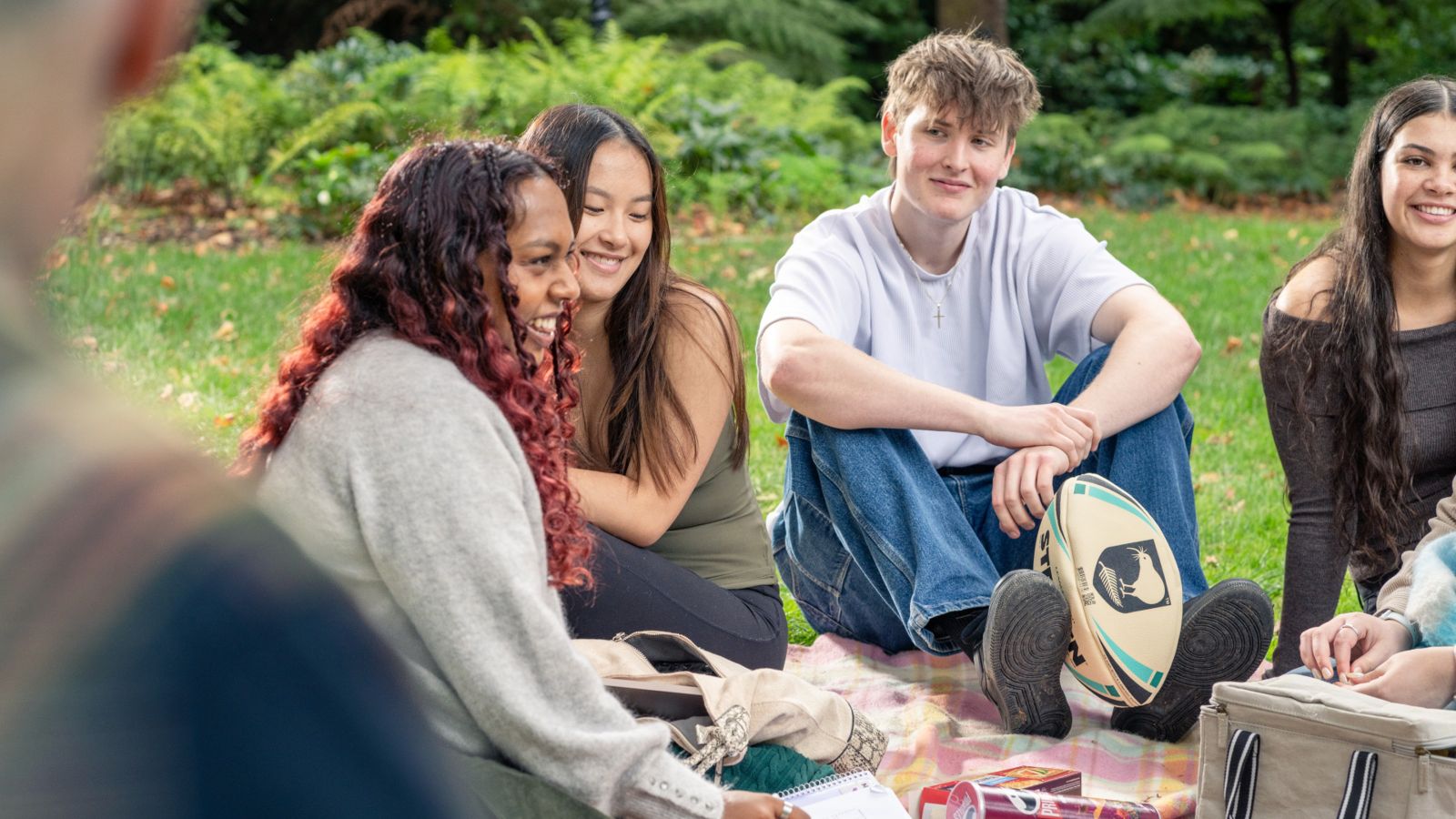 Young people sitting on a picnic mat in the gardens and talking together, one is holding a rugby ball