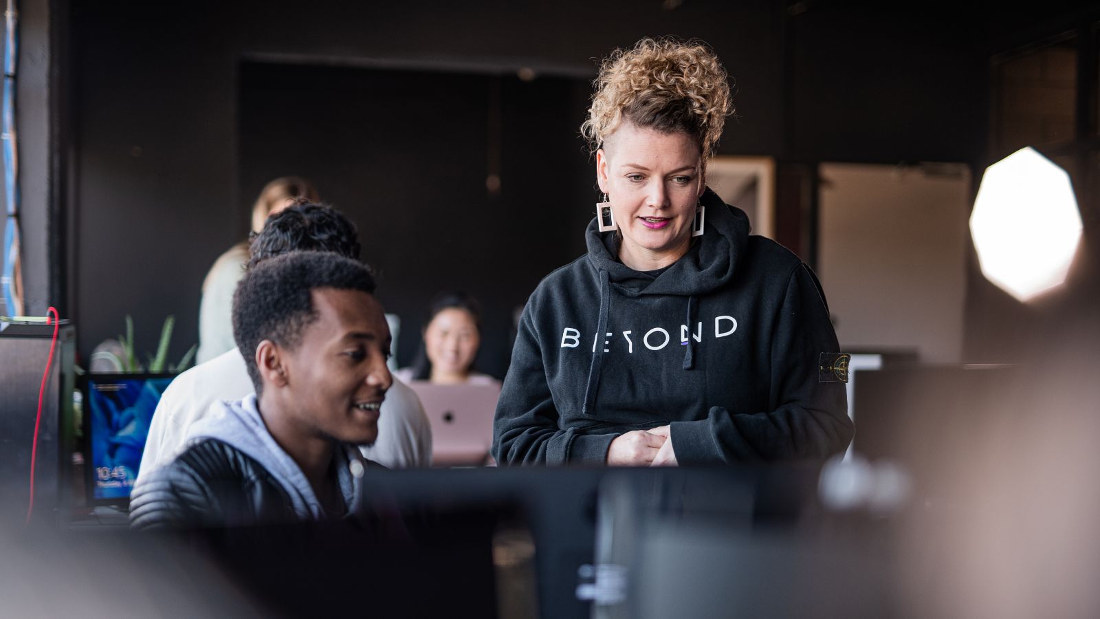 A woman stands beside a student at a screen in the workplace.