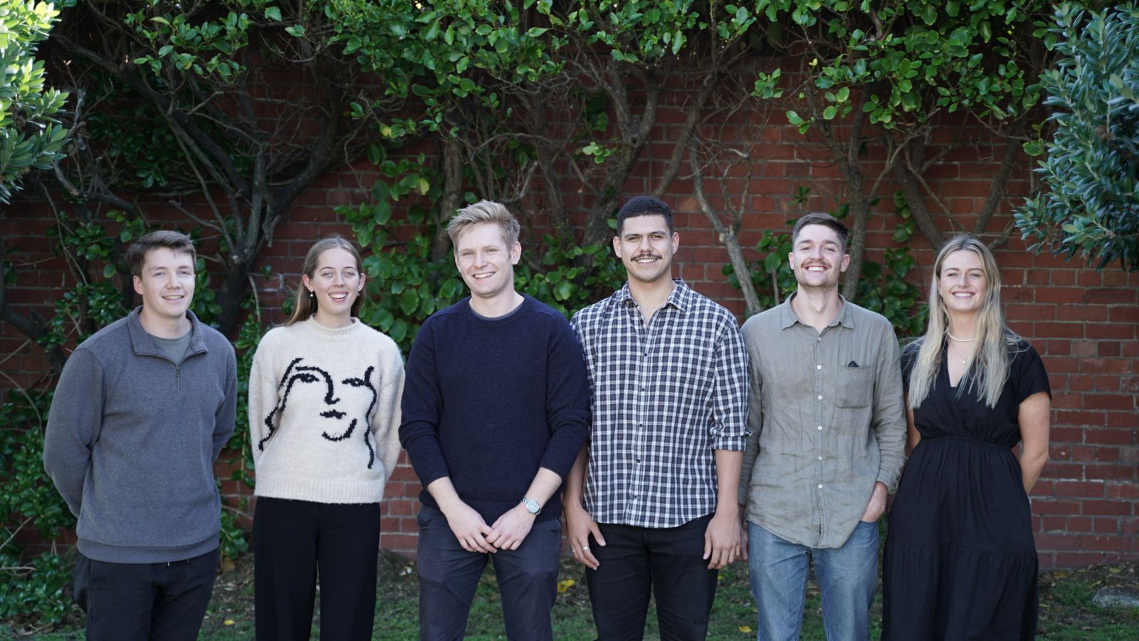 team of six working professionals standing in front of wall with vines.