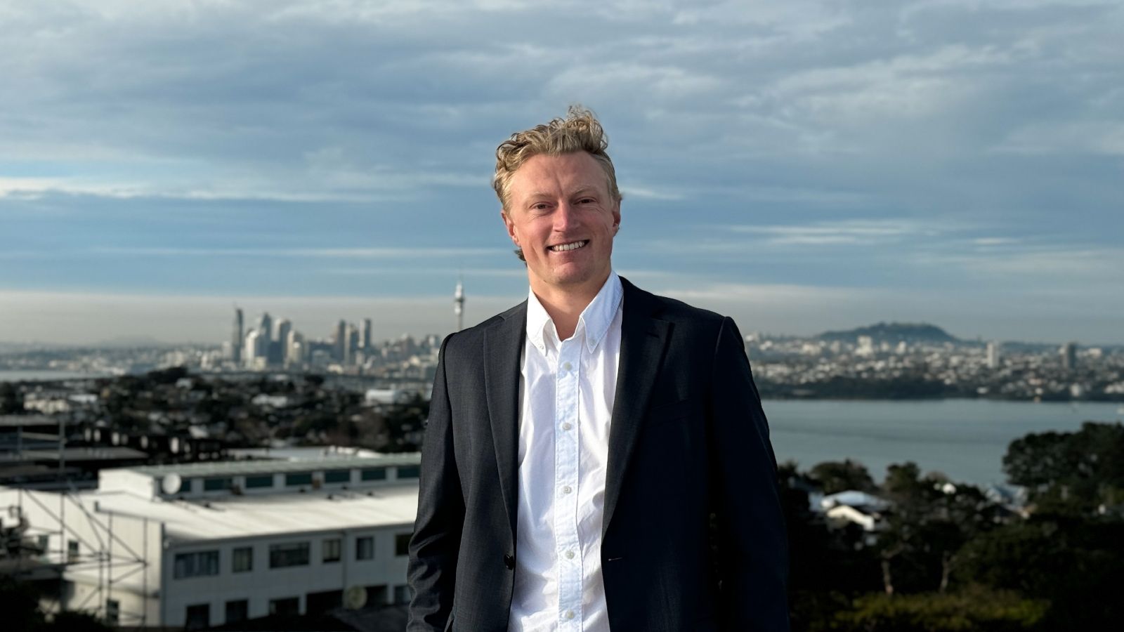 Man in dark suit standing outside with view from Auckland city