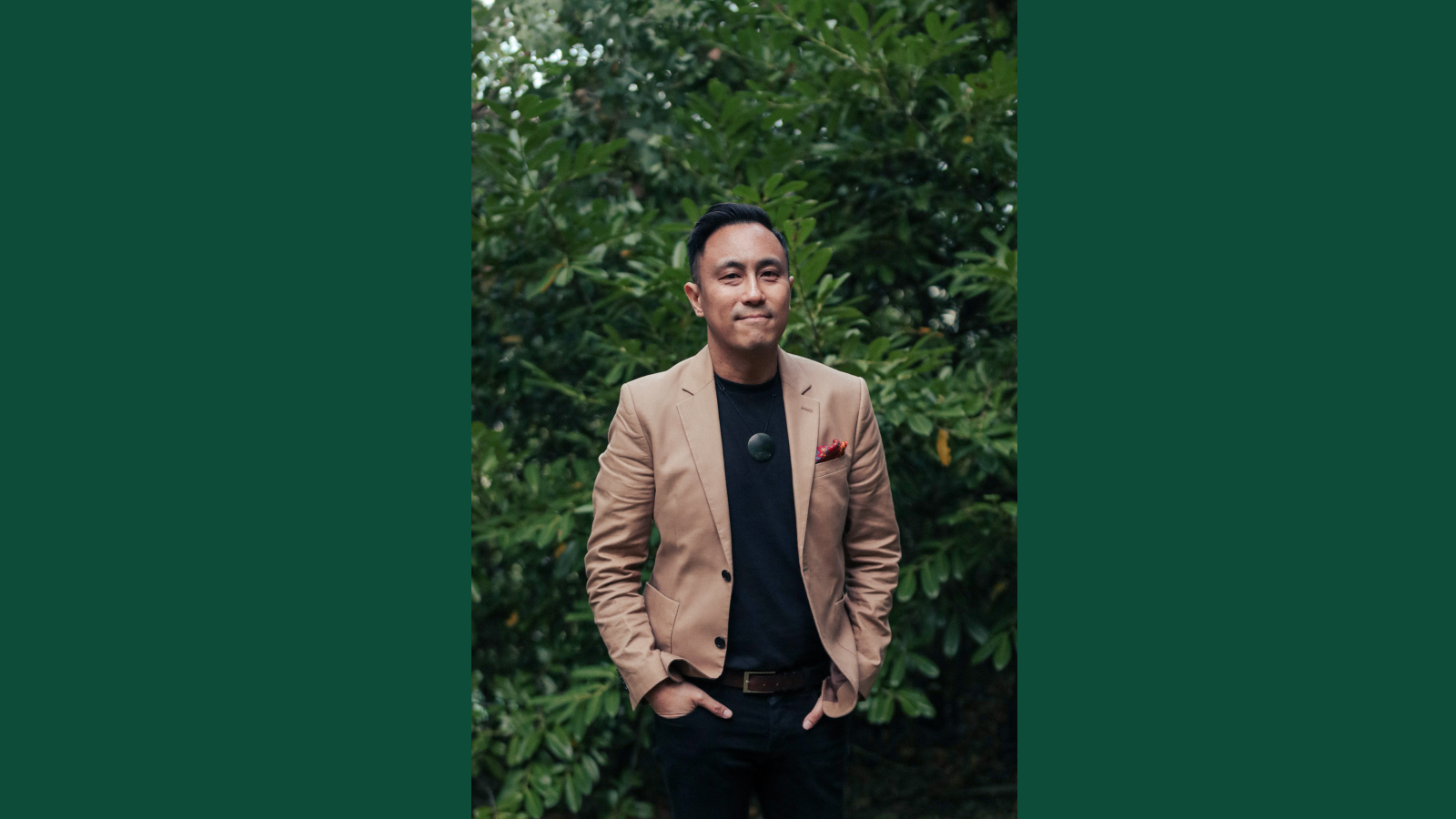 Chris Tse is standing in front of some bushes wearing a blazer with his hands in his pockets and smiling at the camera. 