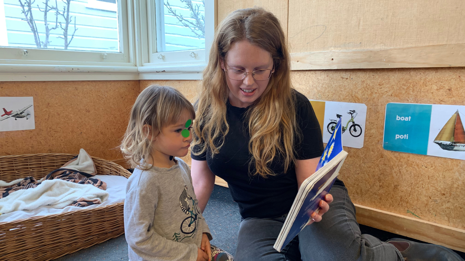 Teacher reading a board book to a child, with bilingual signage around the walls