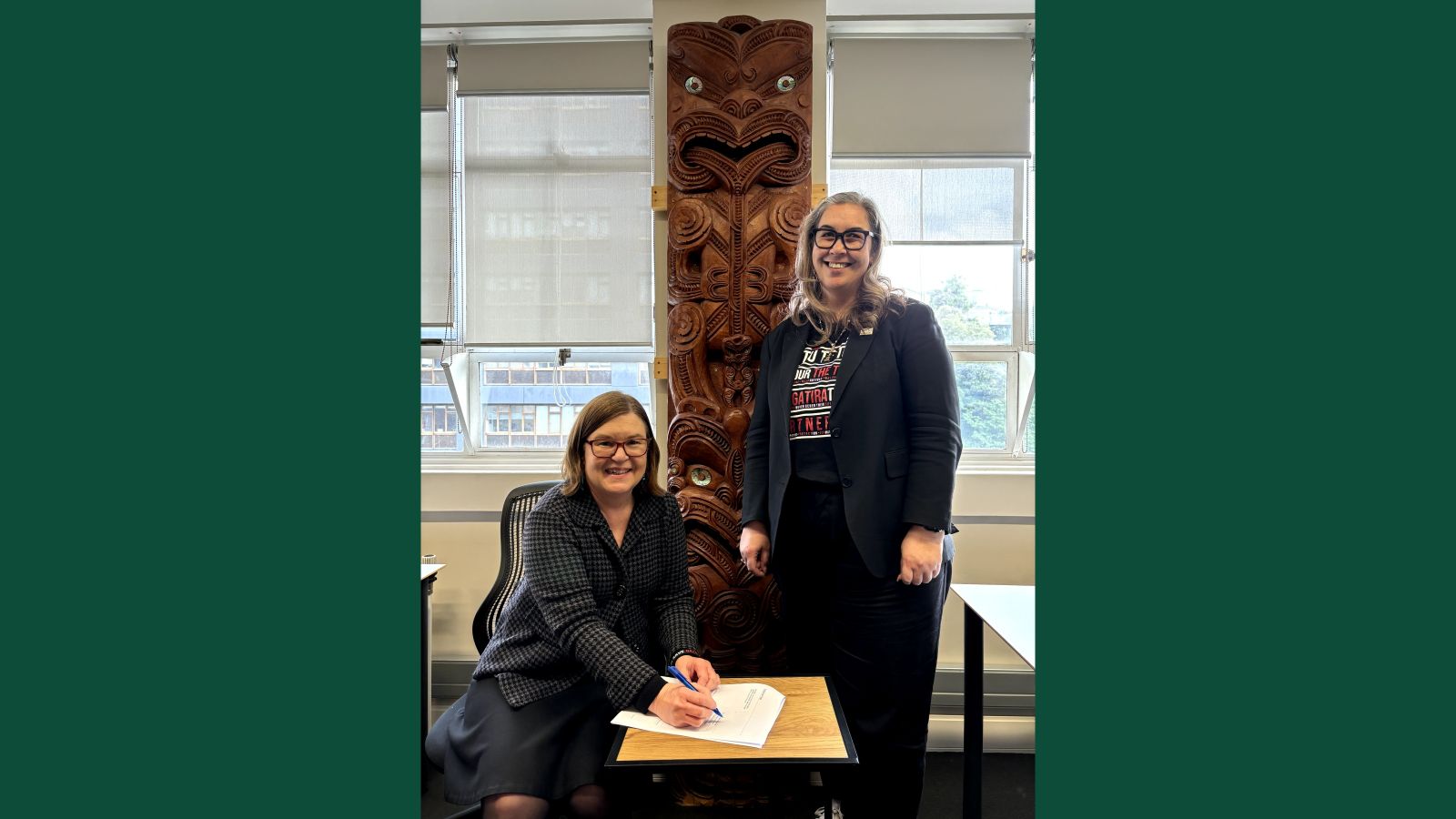 Professor Susan Page is sitting at a table signing a document and Professor Rawinia Higgins is standing next to her. They are both smiling. 