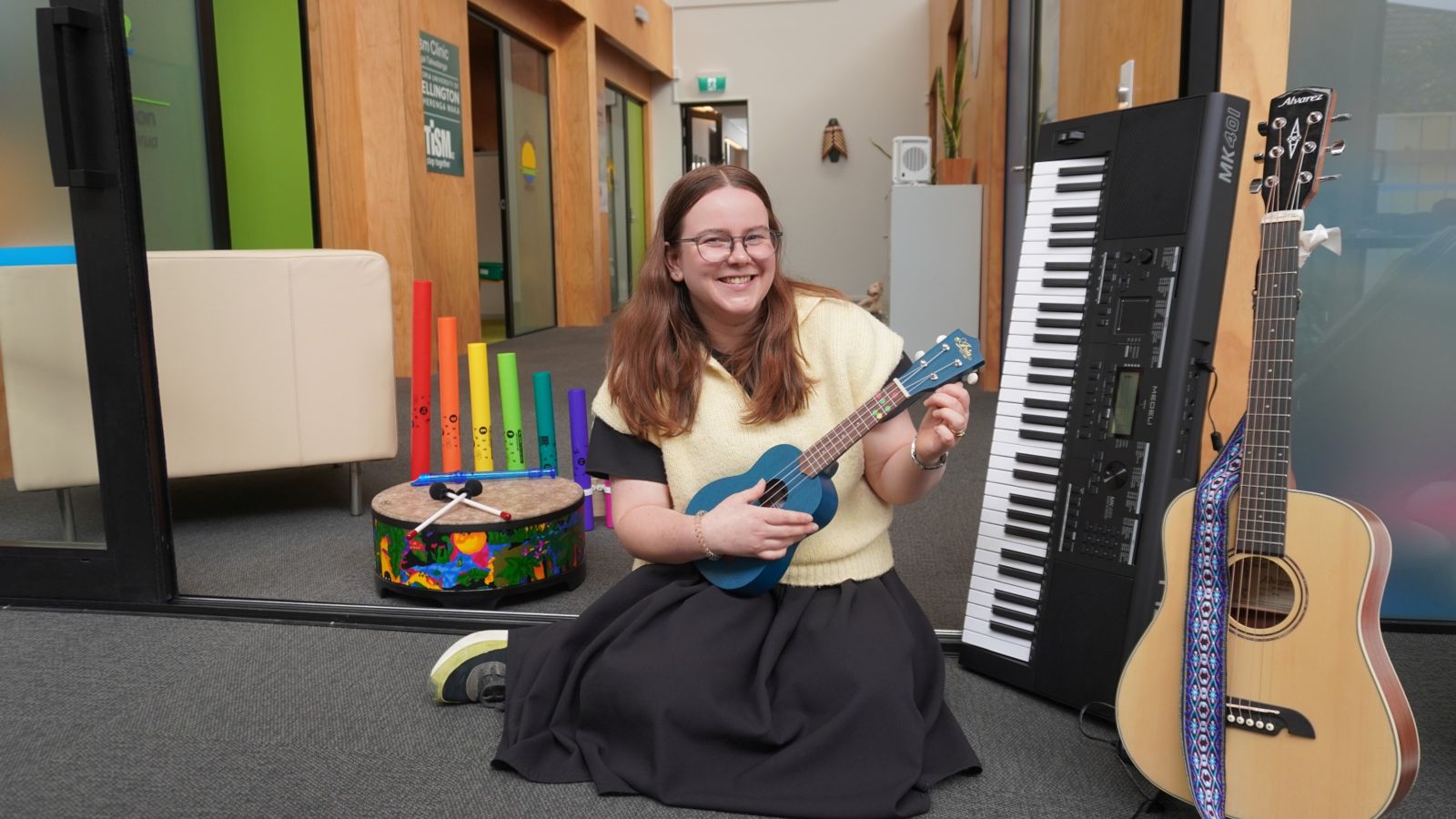 Woman playing ukulele, smiling at camera with other musical instruments around her