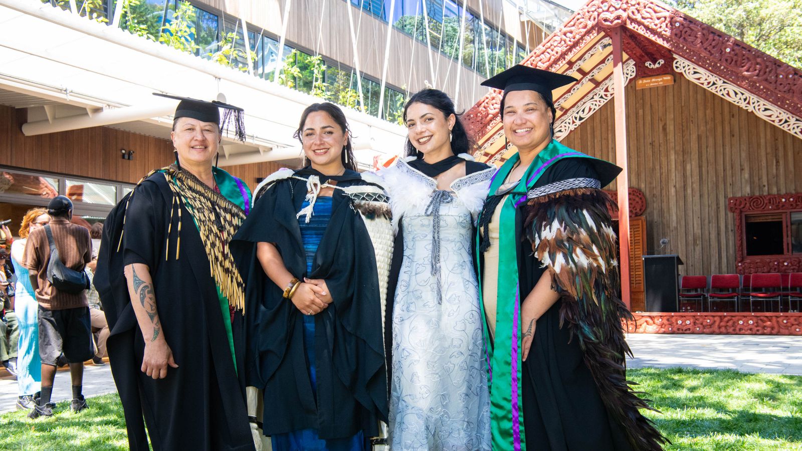 Four wahine Māori are wearing their graduation gear and standing in front of Te Tumu Herenga Waka.