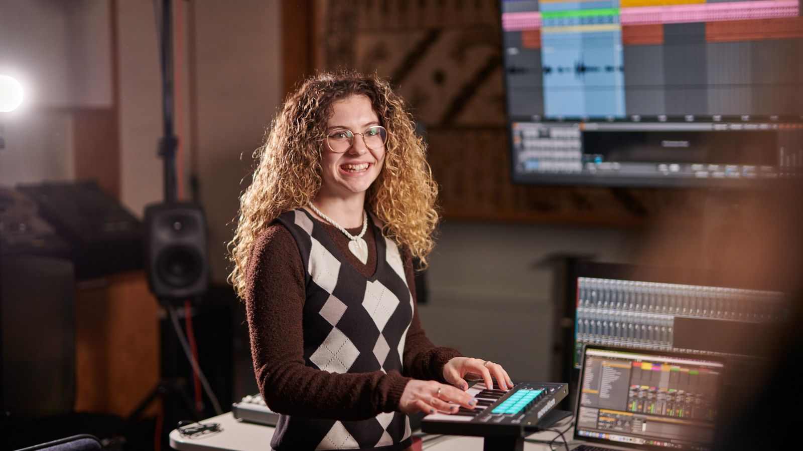 A young woman playing the keyboard and smiling, with screens and equipment in the background