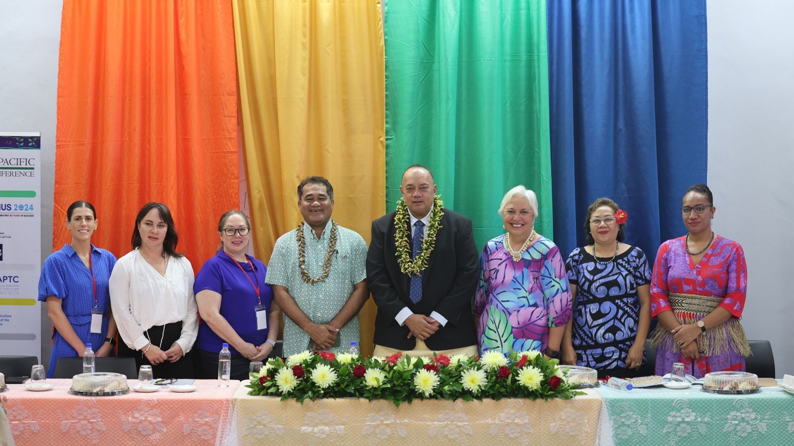 A group of Pacific Island representatives standing together behind a table and smiling, with a rainbow coloured backdrop. 