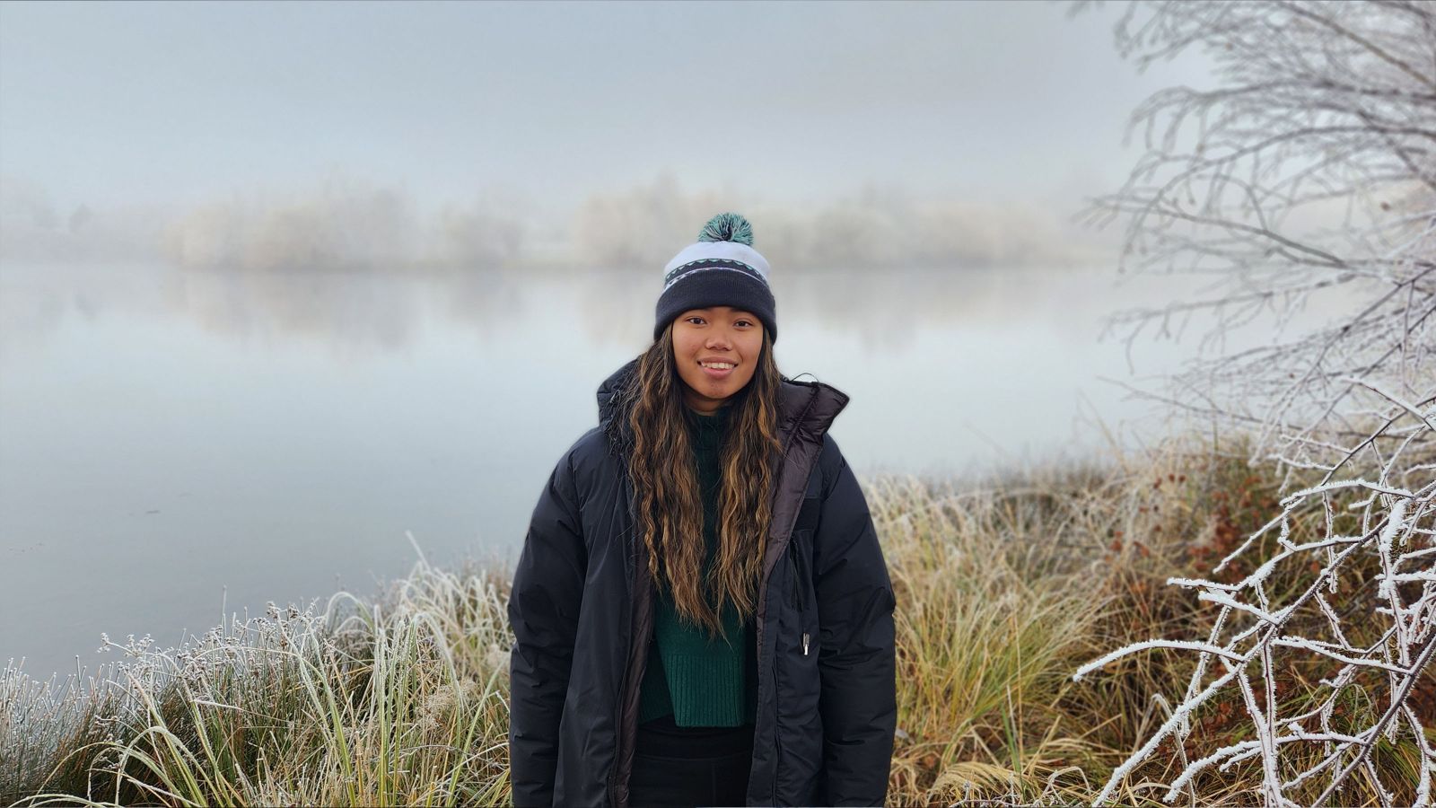 Person in a winter coat and beanie standing near frosty grass and branches by a misty lake on a cold day.